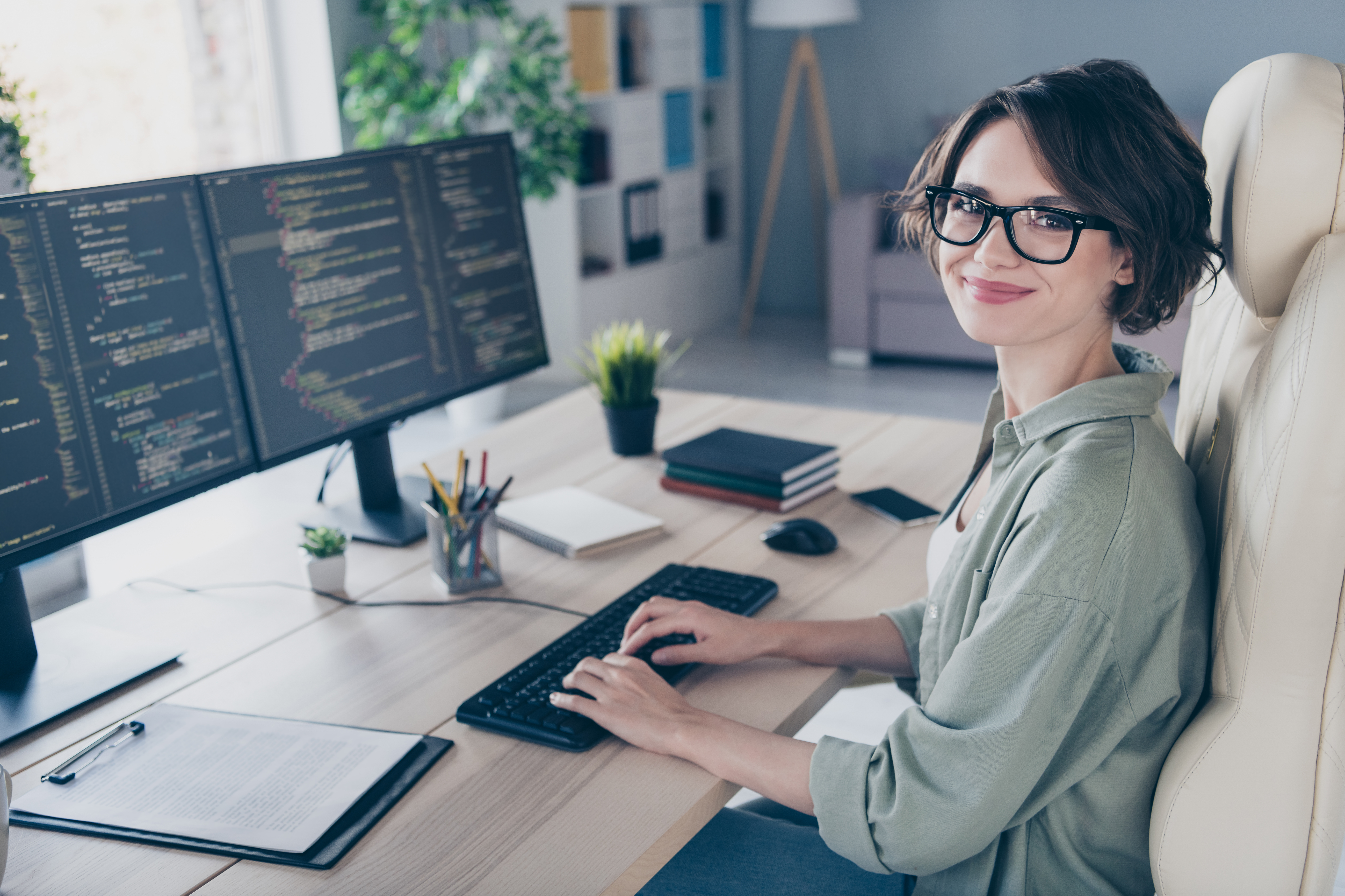 Eine Frau sitzt an einem ergonomisch eingerichteten Schreibtisch mit zwei Monitoren, ihre Hände liegen auf der Tastatur und sie grinst in die Kamera.