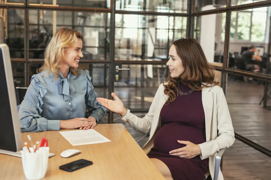 Zwei Frauen unterhalten sich im Büro. Sie sitzen an einem Schreibtisch. Eine der beiden ist schwanger.
